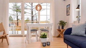 A modern living room with a wooden coffee table, white dining table with chairs, a blue sofa, and a large window with a view of trees. - 4 Star Holiday Home in Laeso (Læsø)