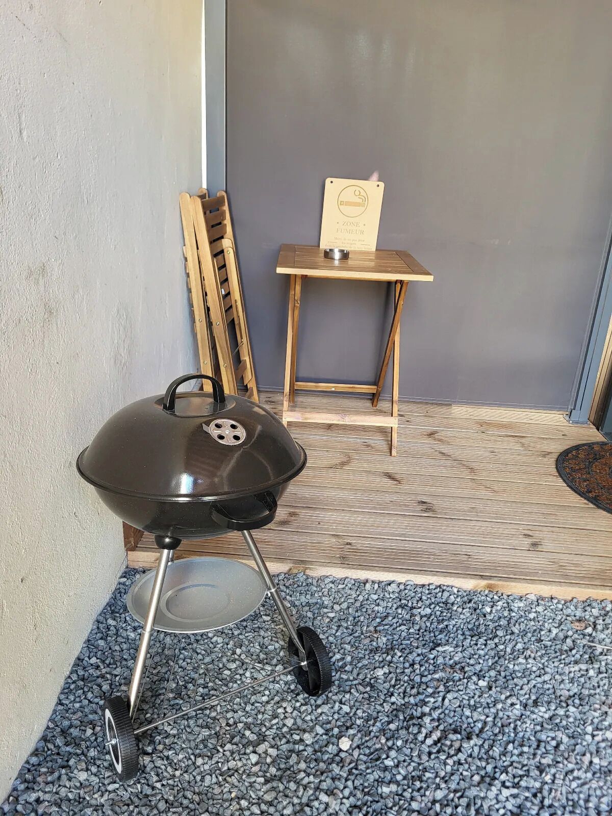 A portable grill with wheels, a wooden chair, and a small table with a sign in a corner.