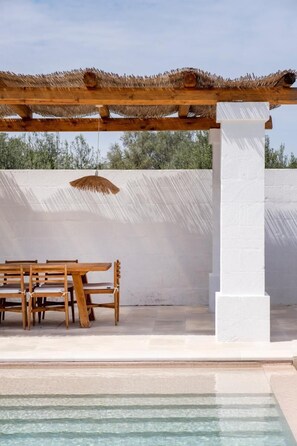 An outdoor seating area with a wooden table and chairs, a thatched roof pergola, and a white wall in the background. - Luxury Trullo with Infinity Pool & Design (Ostuni)