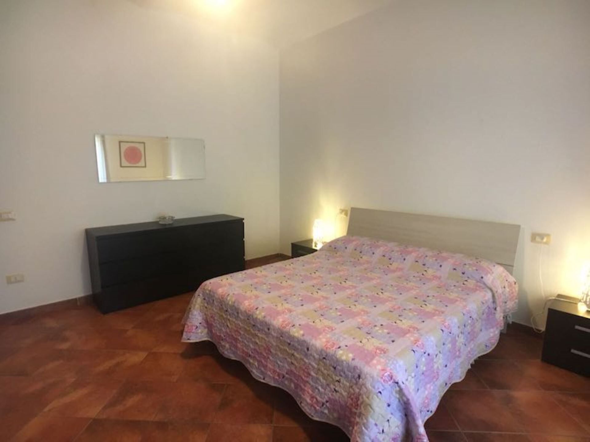 Een slaapkamer met een bed, een dressoir, een nachtkastje en een wandlamp.