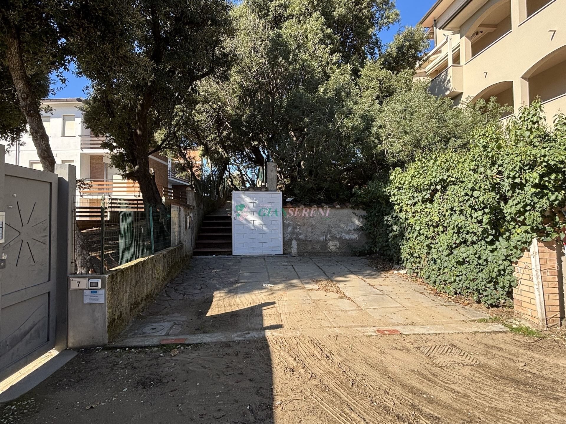 A narrow alleyway with a gate, a staircase leading to a building, and a hedge on the right.