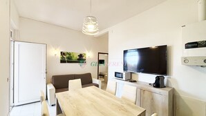A modern living room with a wooden dining table, white sofas, a flat-screen TV, and a ceiling light fixture.
