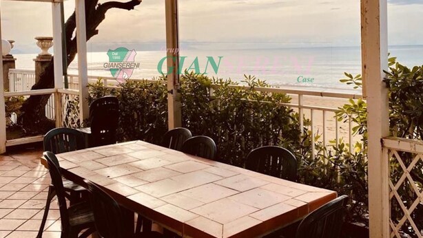 A covered outdoor dining area with a view of the sea, featuring a table and chairs, a pergola, and a decorative tree.