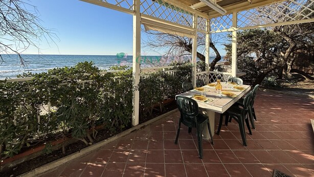 An outdoor dining area with a view of the sea, covered by a pergola with a lattice design, and surrounded by greenery.