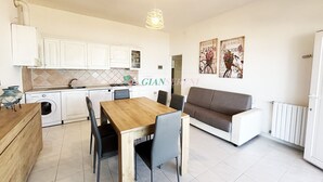 A modern kitchen with white cabinets, a dining table with chairs, and a sofa. There are two wall-mounted light fixtures and a radiator.