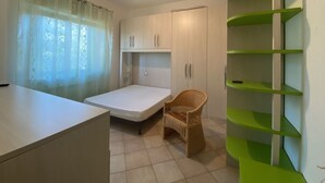 A small, compact room with a single bed, a chair, a desk, and a green shelving unit.