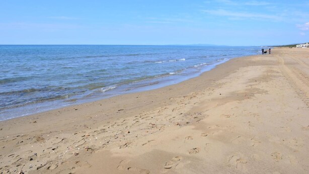 Een breed zandstrand met zachte golven, een heldere lucht en enkele mensen in de verte.