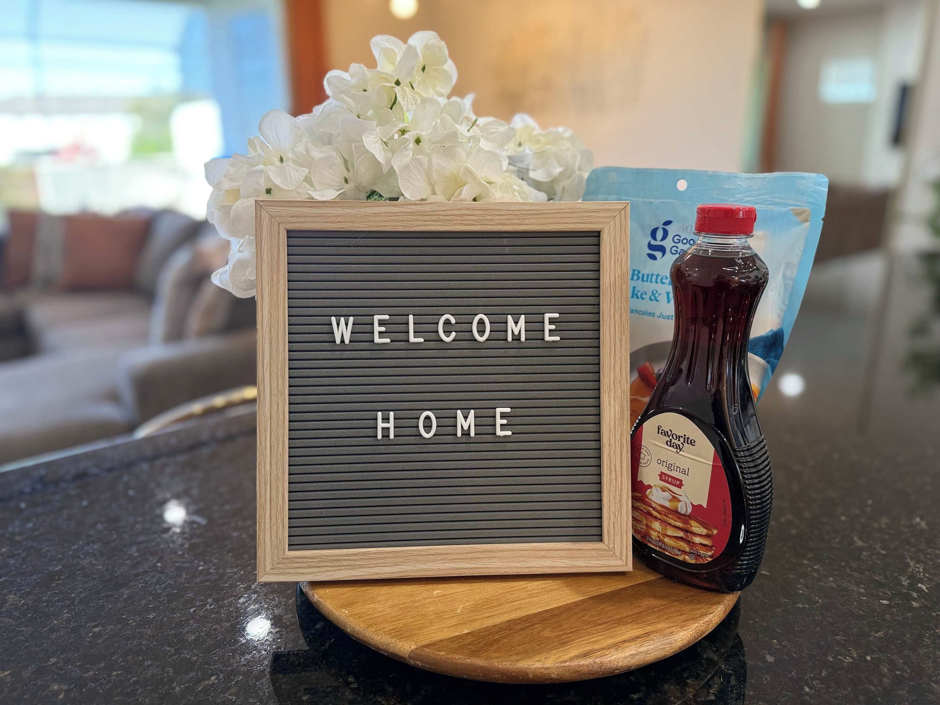 A welcome sign with a message, a bottle of syrup, and a bag of chips on a counter.