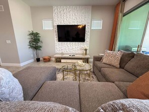 A modern living room with a sectional sofa, a flat-screen TV, and a decorative wall panel. - Escape to a 4BR Villa w/ Arcade, Pool & Mini Golf (Davenport)