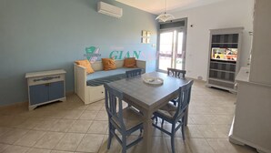 A dining area with a table and chairs, a sofa with cushions, a cabinet, and a television displaying a news broadcast. - TWO-ROOM APARTMENT SLEEPS 4 IN THE LA PIANA AREA (San Vincenzo)