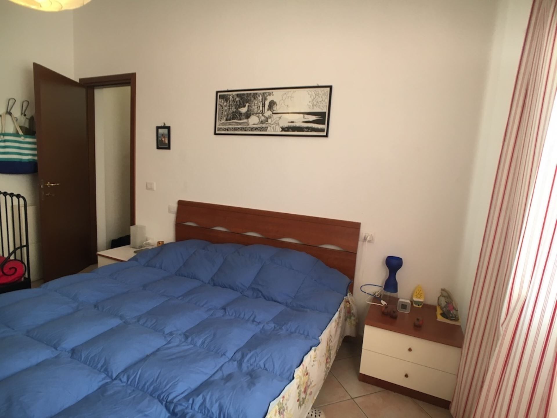 Quarto com cama, mesa de cabeceira, quadro na parede e porta.