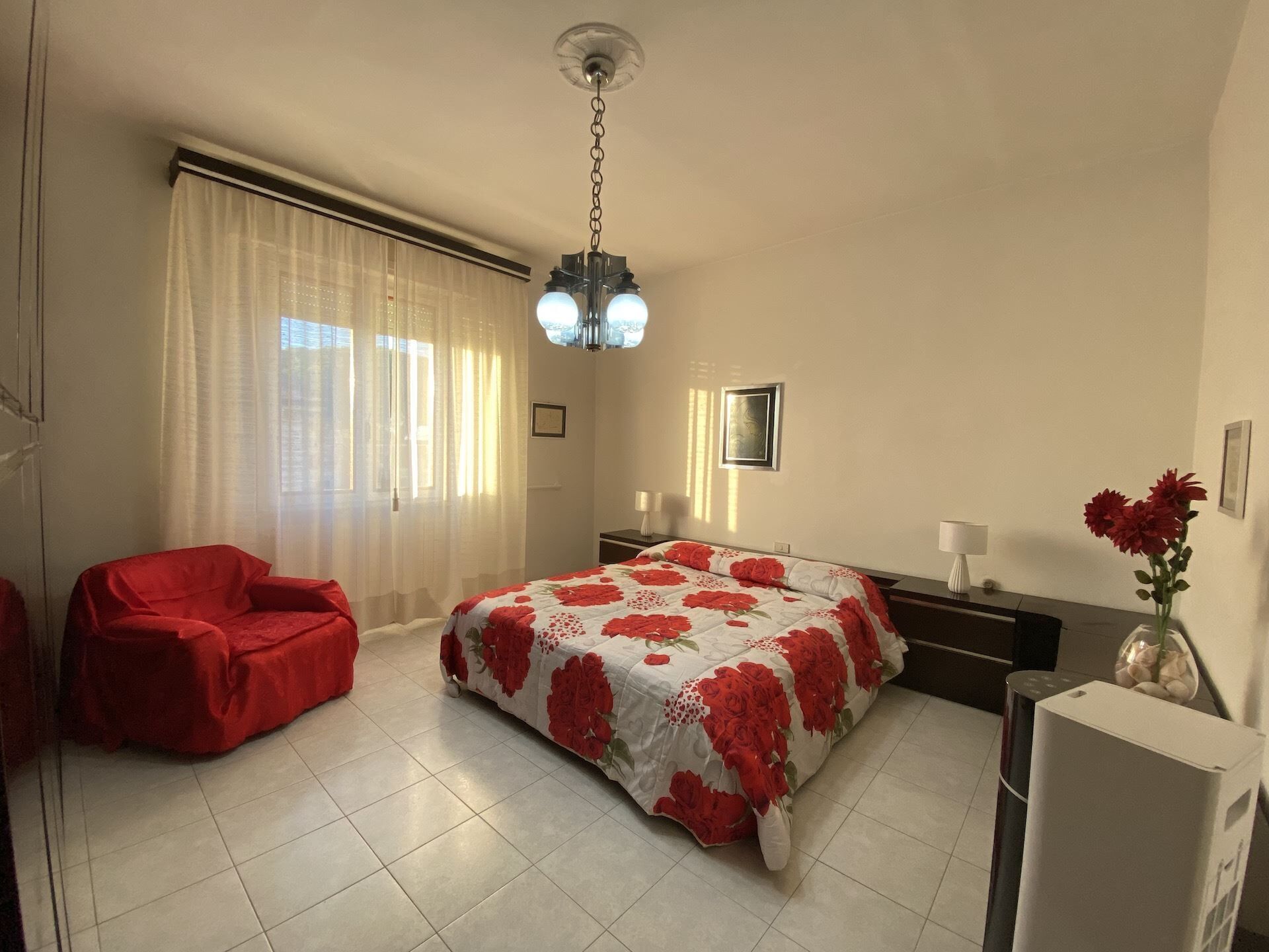 Ein Schlafzimmer mit einem Bett, einem roten Sessel, einem Schreibtisch, einer Lampe und einer Vase mit Blumen.