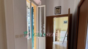 Un pasillo con suelo de baldosas, una puerta de madera y vista a otra habitación con suelo de cerámica y un cuadro en la pared.