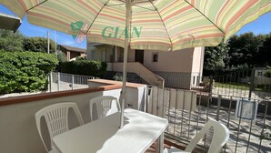 A terrace with a striped umbrella, white plastic chairs, and a table. - Apartment in the Bagno Venere area (San Vincenzo)