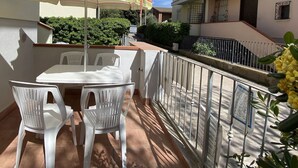 Un balcon avec une table et des chaises blanches, une rambarde en métal et une vue sur la rue et les bâtiments voisins.