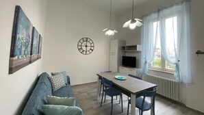 A modern living room with a sofa, dining table, and chairs. There is a clock on the wall and a window with curtains. - 5 bed places with parking space - ROSS2S (San Vincenzo)