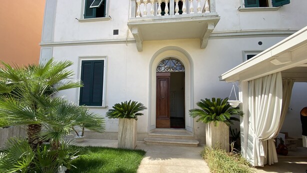 A two-story building with a central entrance, a balcony with white balustrades, and a small garden area with palm trees and a pathway. - 5 bed places with parking space - ROSS2S (San Vincenzo)