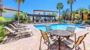 Un espace aménagé au bord de la piscine, avec des chaises longues et une table, entouré de palmiers et d’un bâtiment doté d’un toit en tuiles.