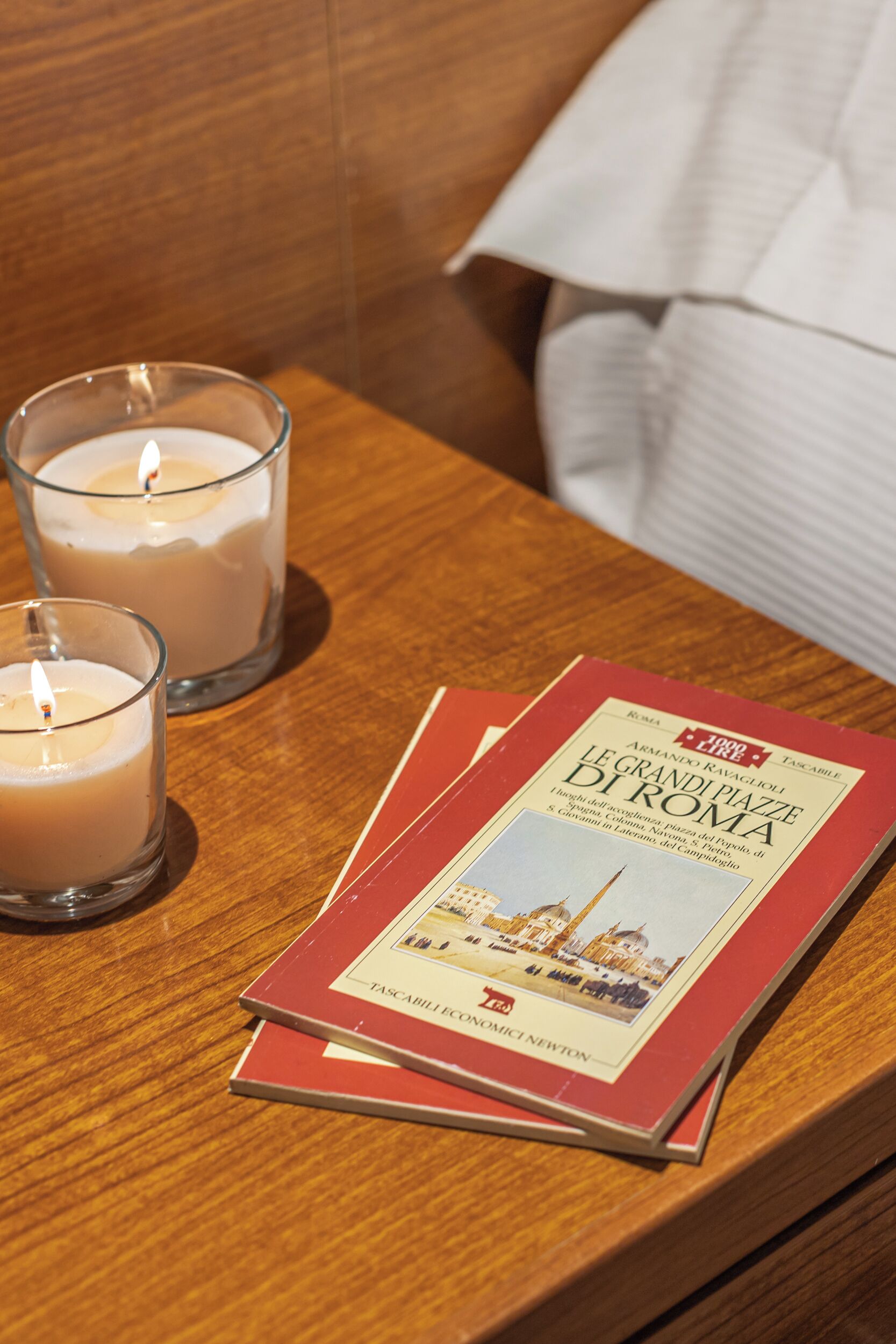 Zwei Kerzen auf einem Holztisch, ein Buch mit dem Titel "Le Grandi Plais di Roma" und ein weißes Kissen im Hintergrund.