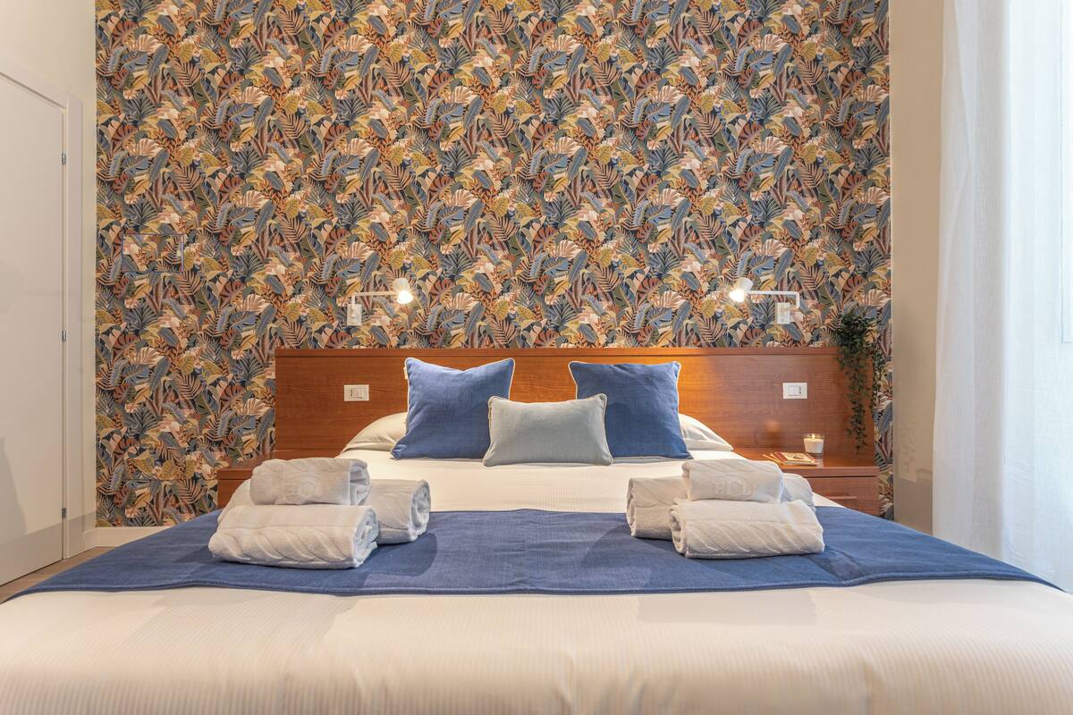 A hotel room with a large bed, two pillows, a nightstand, and a wall with a floral pattern.