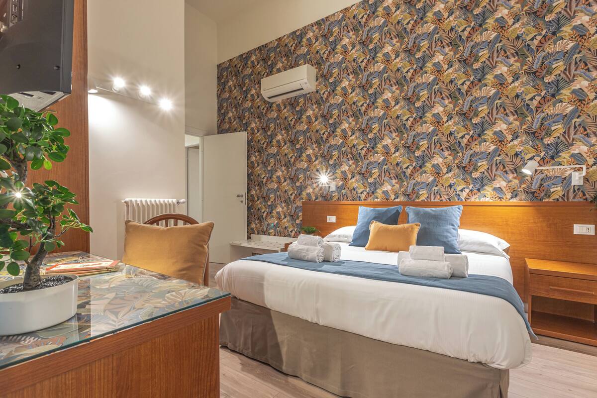 A hotel room with a bed, a nightstand, a bedside table, a potted plant, and a patterned wall.