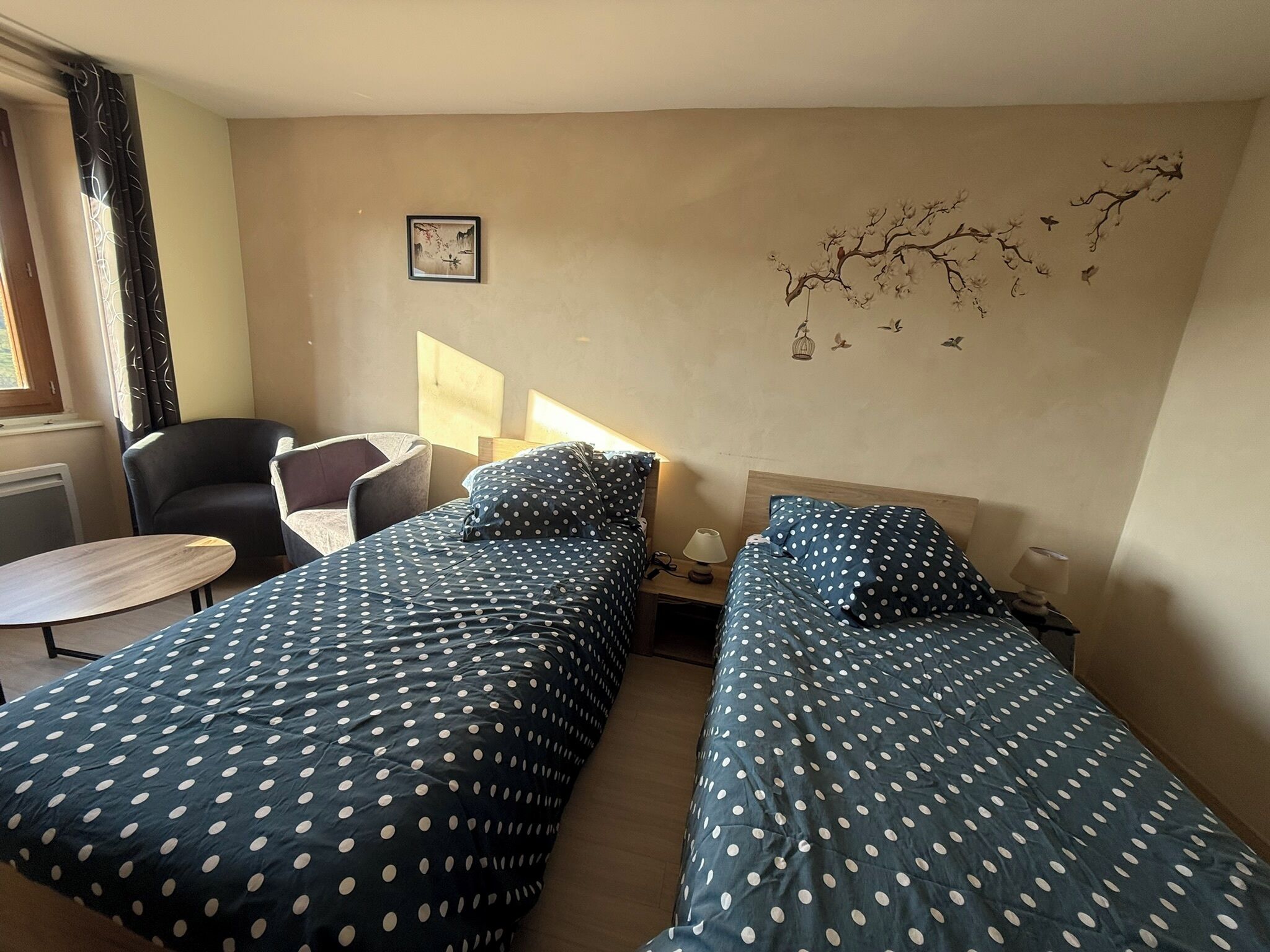 A hotel room with two single beds, a small table, a chair, a nightstand, and a wall decoration.