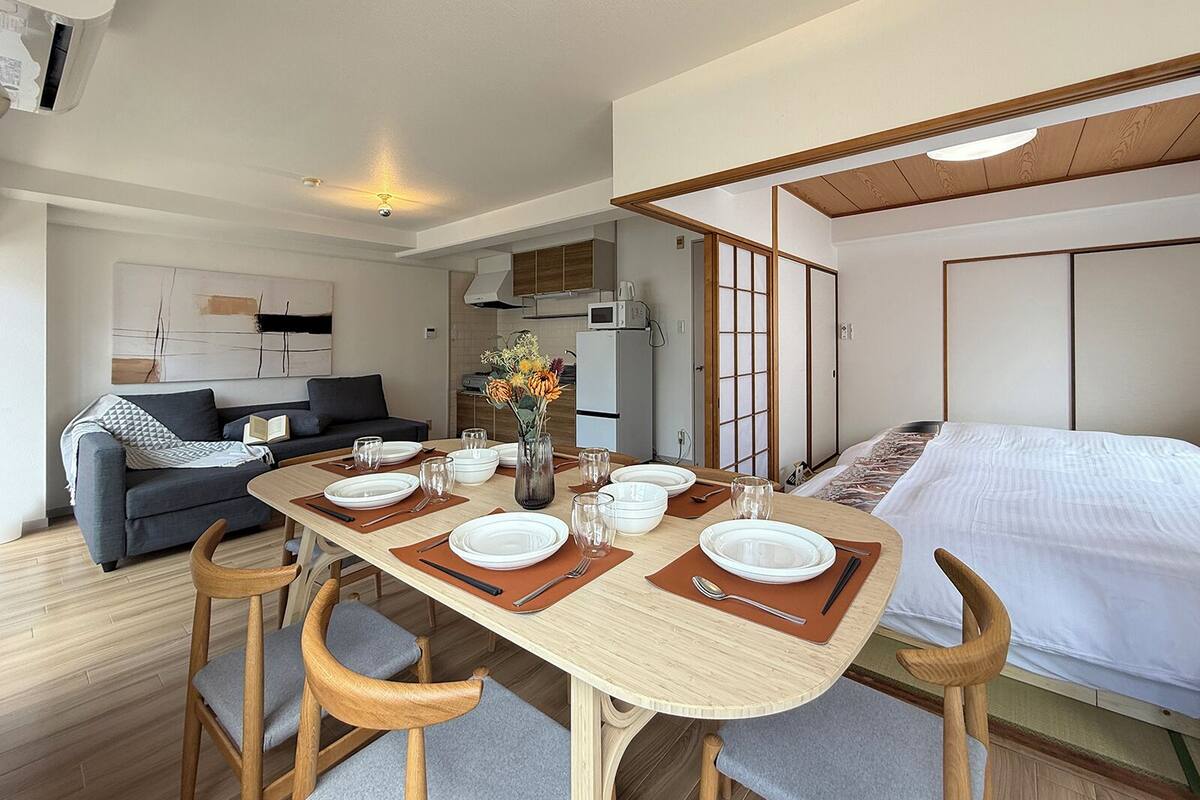 A modern, compact living space with a dining area set for four, a sofa, a kitchen, and a bedroom with a sliding door.