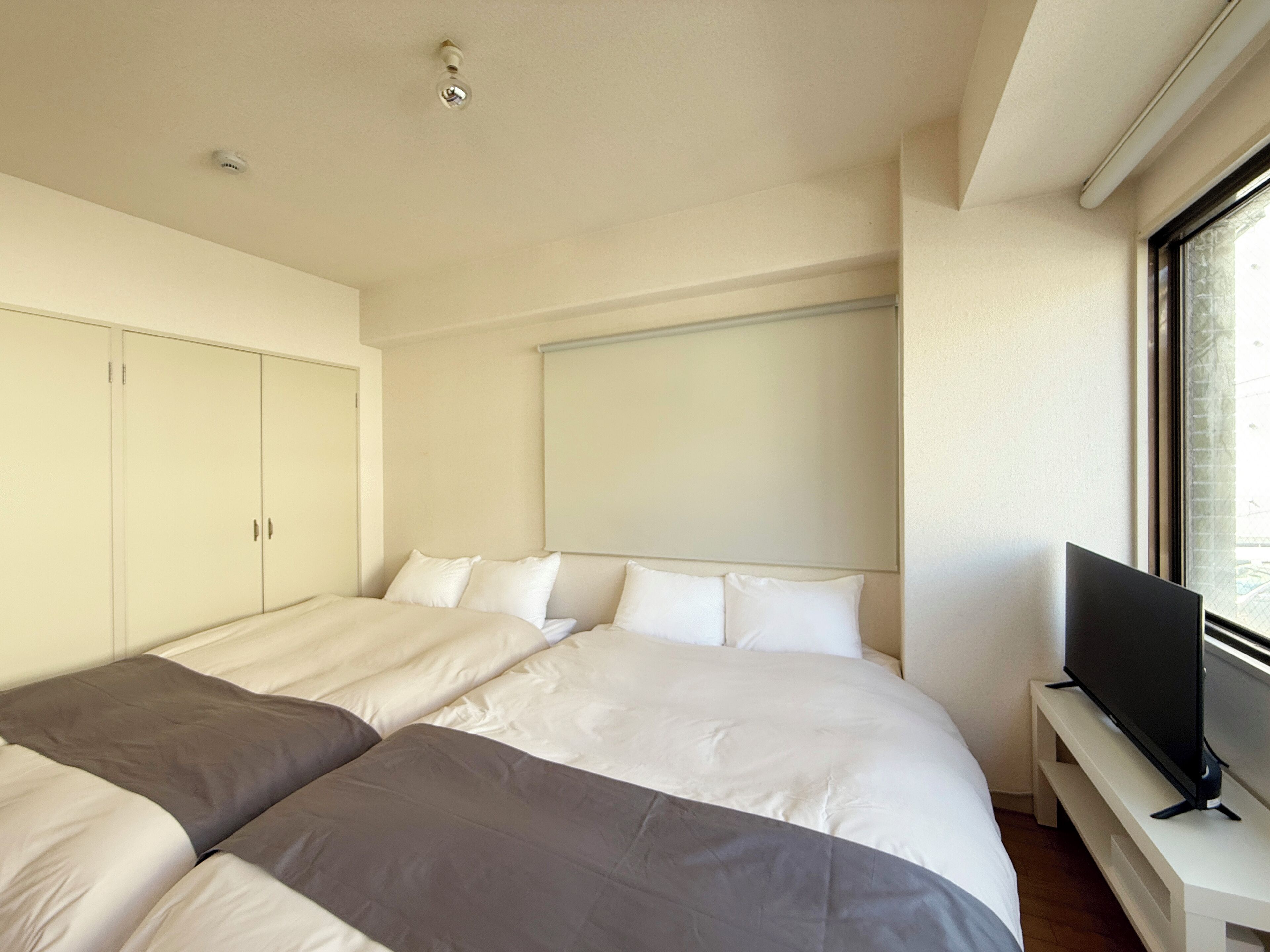 A modern hotel room with two single beds, a flat-screen TV on a stand, and a large window providing a view of the outside.