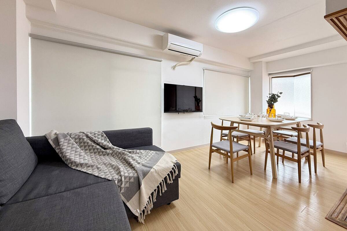 A modern living room with a grey sofa, a dining table with chairs, a flat-screen TV mounted on the wall, and an air conditioning unit.
