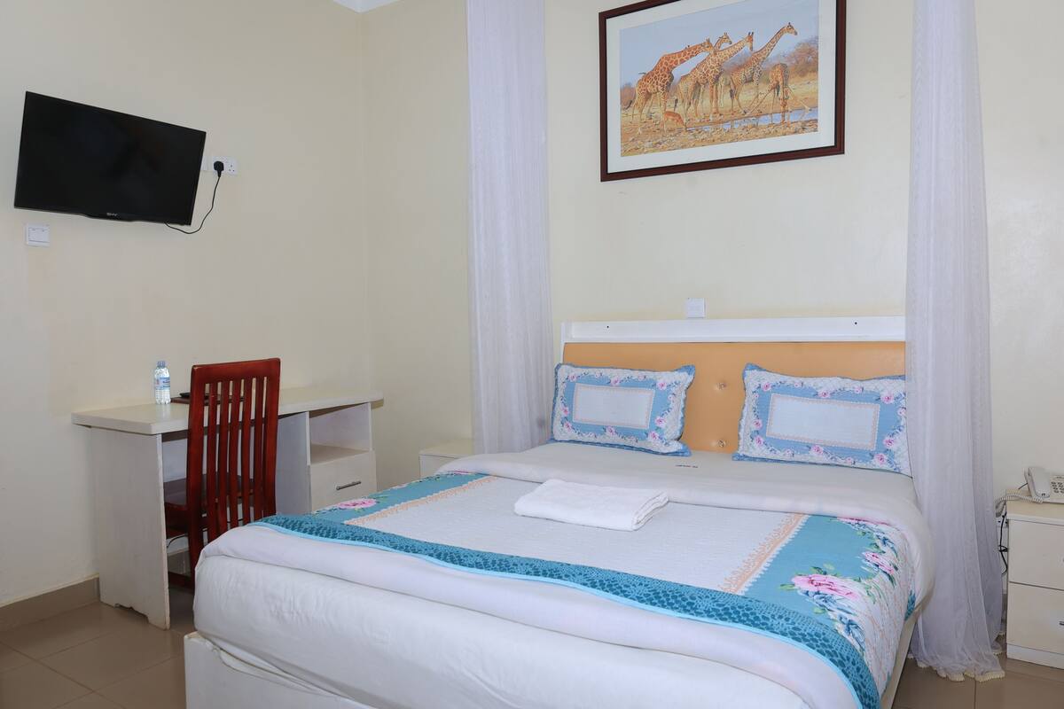 A bedroom with a bed, a desk, a chair, a television mounted on the wall, and a painting of giraffes.