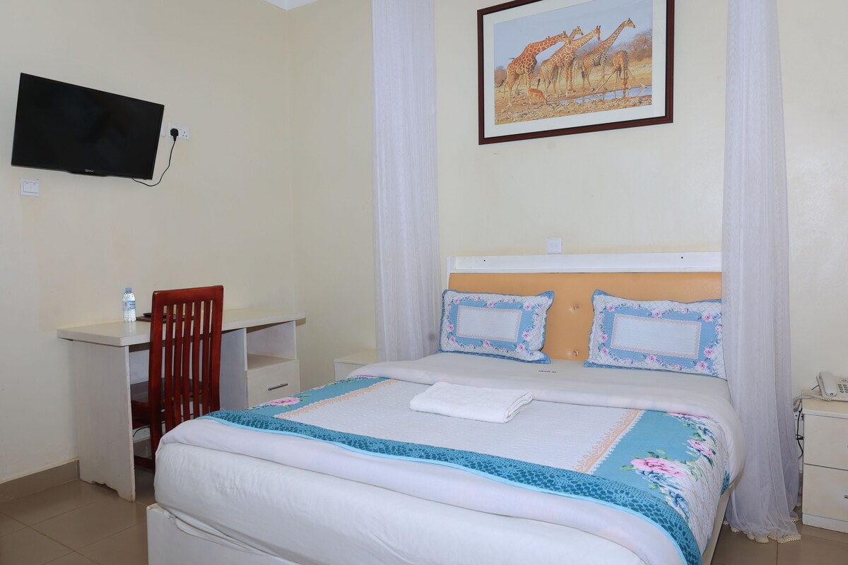 A bedroom with a bed, a desk, a chair, a television mounted on the wall, and a painting of giraffes.