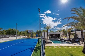 Uma quadra de tênis, uma piscina e uma área de estar coberta com palmeiras.