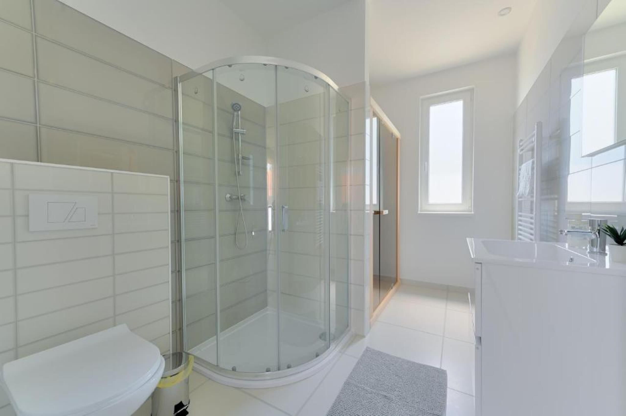 A modern bathroom with a glass-enclosed shower, a white sink, and a large window providing natural light.