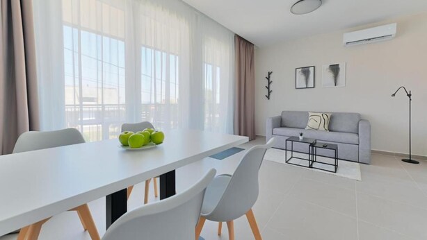 Een moderne woonkamer met een witte bank, een eettafel met stoelen en een groot raam met dunne gordijnen.