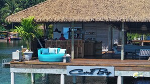 Uma cabana com telhado de palha, área de bar, sofá turquesa e uma placa com a inscrição 'Cool Bay'.