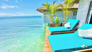 Um bangalô à beira-mar com um deck de madeira, duas poltronas azuis-turquesa e uma estrutura com telhado de palha.