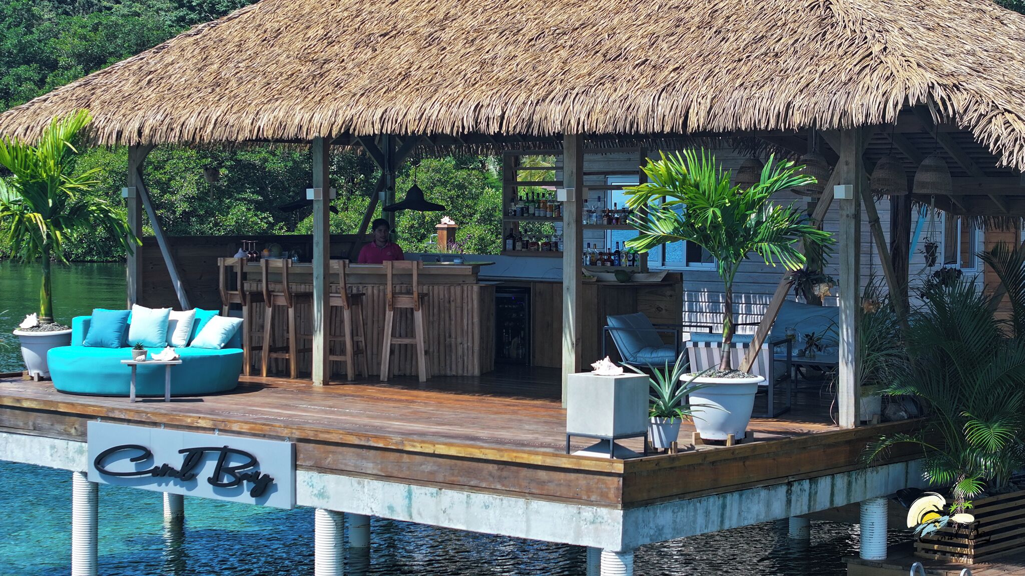 A thatched-roof overwater bungalow with a wooden deck, a bar area, and seating.