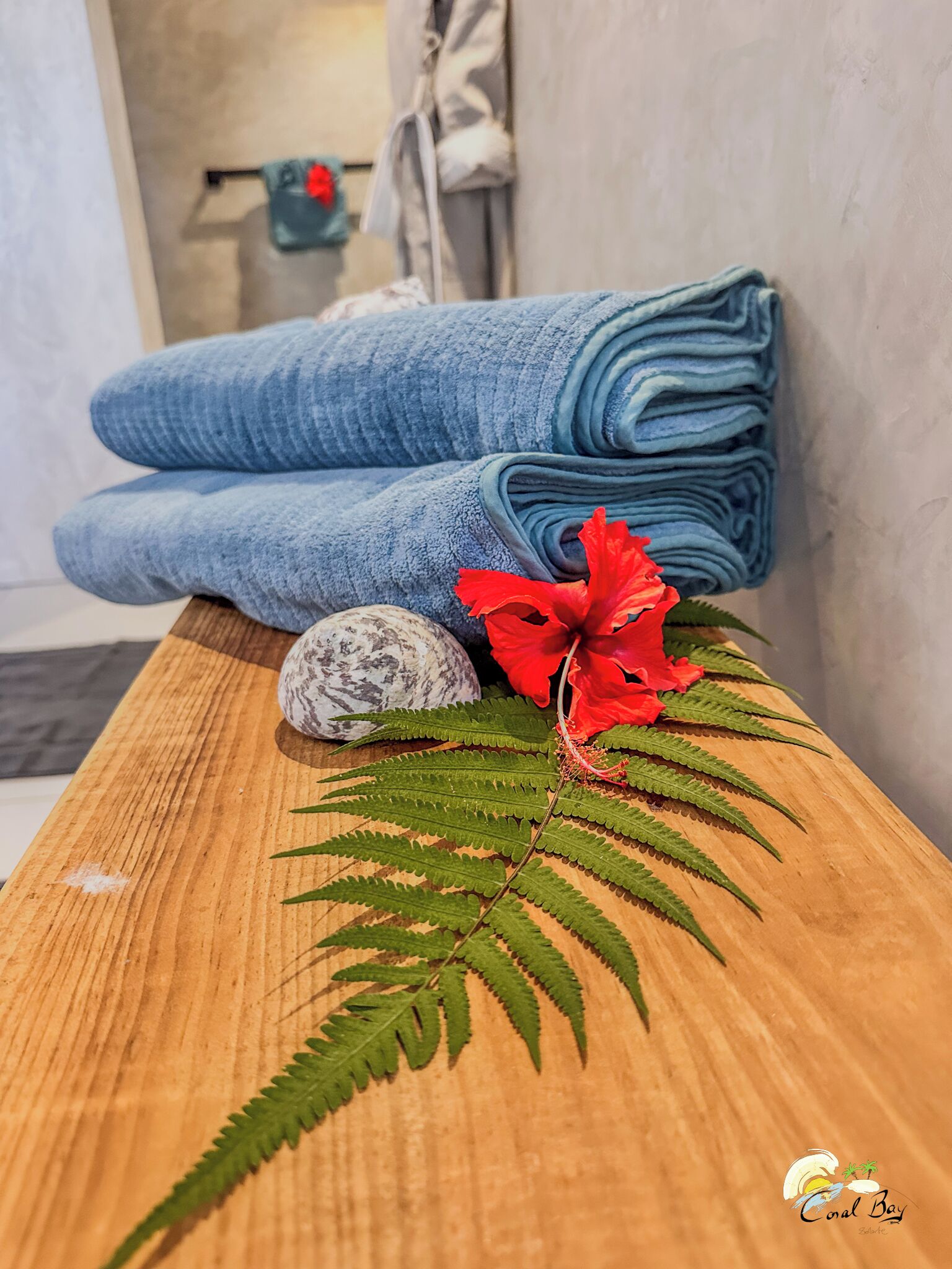 Ein Holztisch mit einem blauen Frotteetuch, einer roten Hibiskusblüte, einem Stein und einem Farnblatt.
