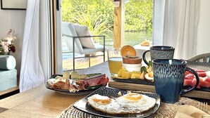 Mesa de café da manhã com pratos de comida, uma caneca e um copo de suco, com vista para um jardim exuberante.