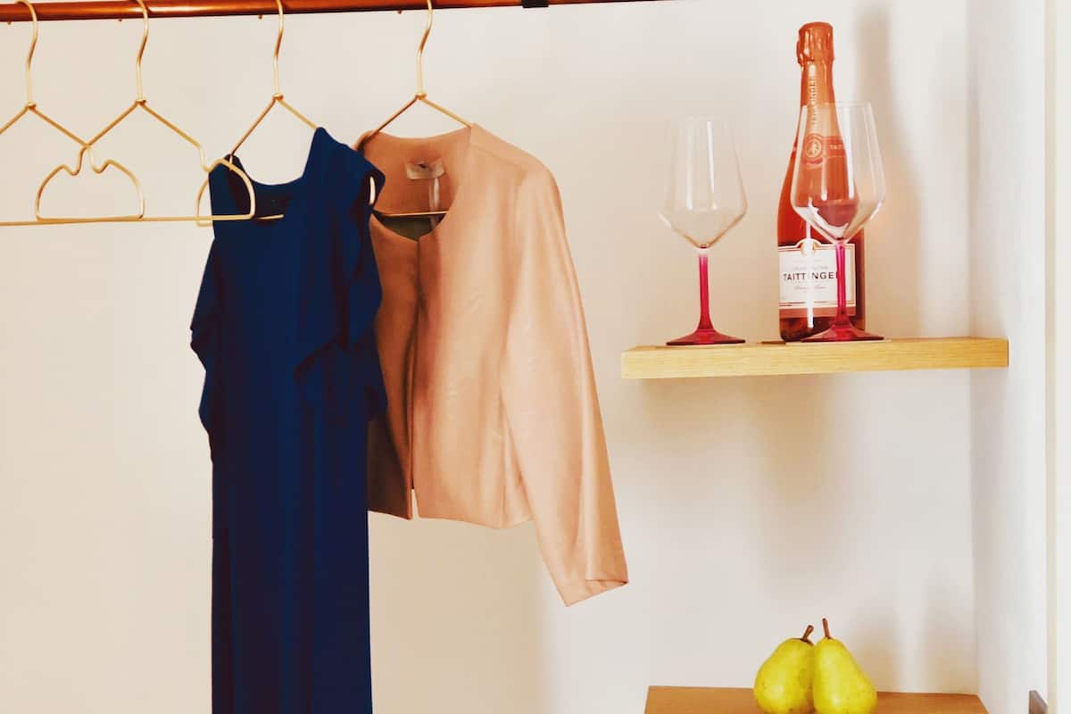A room with a clothing rack holding a blue dress and a beige jacket, a wine bottle and glass on a shelf, and a wooden shelf with two pears.