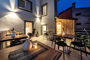 Un área de comedor al aire libre con mesas y sillas, una pérgola de madera y vistas a la ciudad al anochecer.