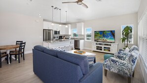 A modern living room with a blue sofa, a dining table with chairs, a kitchen area with white cabinets, a ceiling fan, and a painting on the wall. - ☀️ Private Pool - Short Walk to Beach - Betty's Beach House ☀️ (Fort Myers Beach)