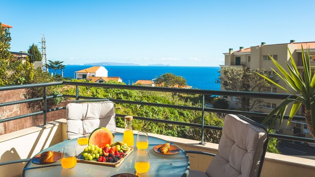 A balcony with a table set for breakfast, including fruits and juice, overlooking a coastal view with buildings and the sea. - Sweet Home Reis Magos by Atlantic Holiday (Caniço)