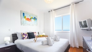A neatly arranged hotel room with a bed, a bedside table with a lamp, a small TV, and a window with curtains. - Sweet Home Reis Magos by Atlantic Holiday (Caniço)