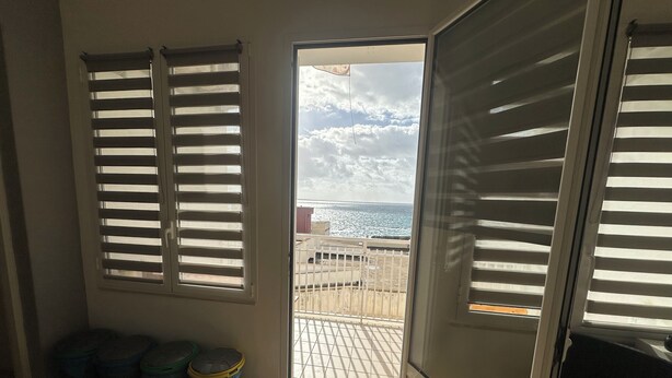 Ein Zimmer mit einer Schiebeglastür, die auf einen Balkon mit Meerblick führt. Zwei Mülleimer und ein Fenster mit Jalousien sind ebenfalls vorhanden.