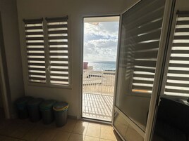 A room with a sliding glass door leading to a balcony with a view of the sea. There are two trash bins and a window with blinds.