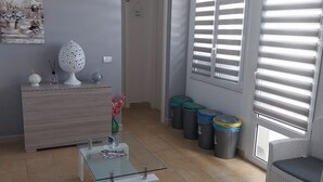 Ein Wohnzimmer mit einer Couch, einem Couchtisch, einem Fernseher und einer Tür zu einem anderen Raum. Mülleimer und ein Fenster mit Jalousien sind ebenfalls vorhanden.