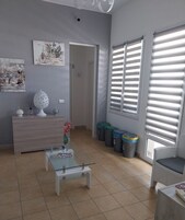 A living room with a sofa, a coffee table, a TV, and a door leading to another room. There are trash bins and a window with blinds.