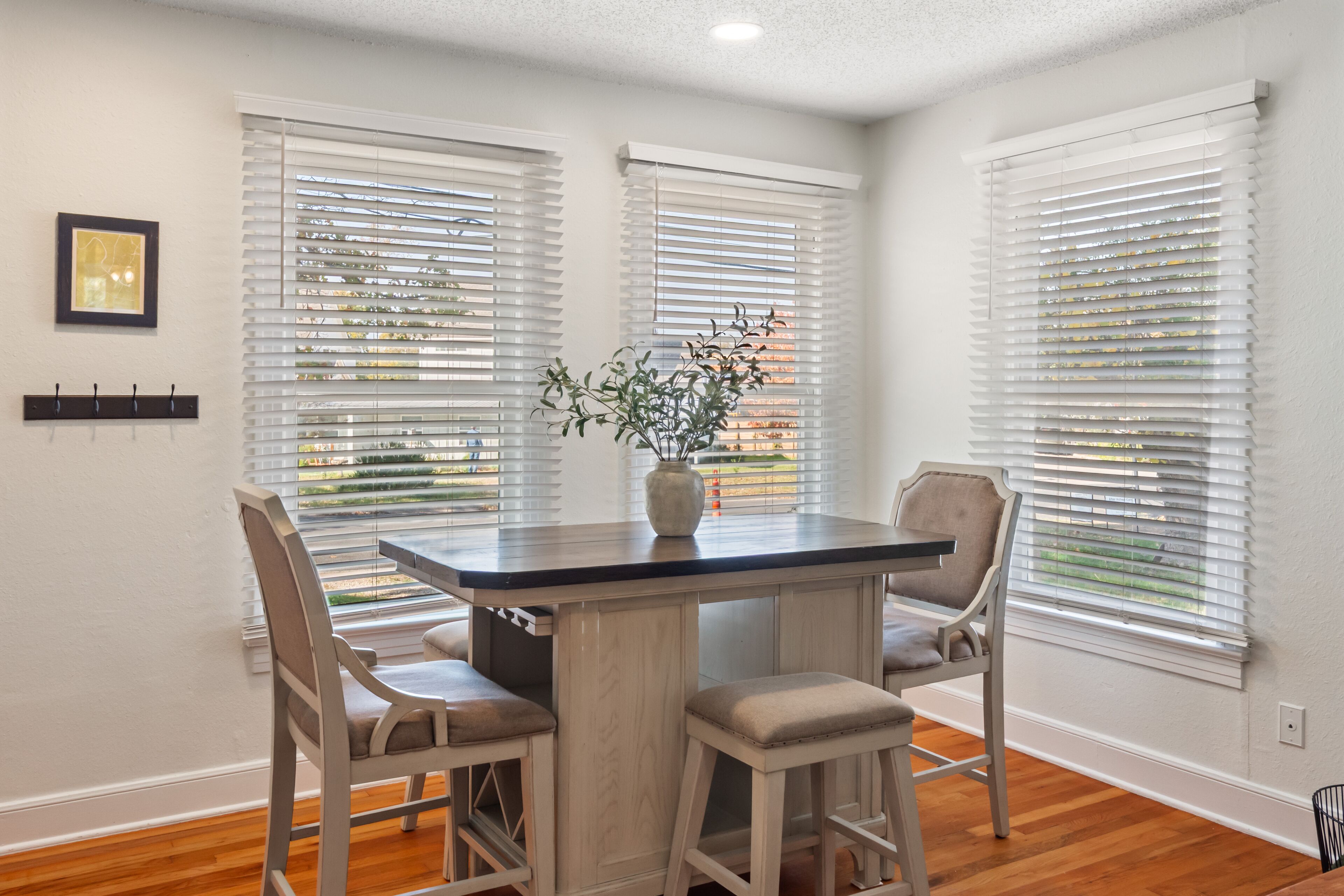 A dining area with a table and four chairs, white walls, and large windows with blinds.