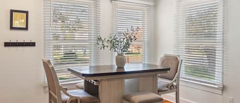 A dining area with a table and four chairs, white walls, and large windows with blinds.